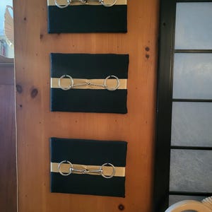 May include: Three rectangular wall art pieces featuring black canvases with a horizontal gold stripe across the center. Each canvas displays a silver-toned metal horse bit attached to the front, showcasing different bit designs including a chain link.