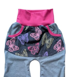 May include: Gray baby pants with a pink waistband and pocket trim. The front panel features a dark gray fabric with pink, purple, and gold butterfly designs. The pants have a high waist and a comfortable fit.
