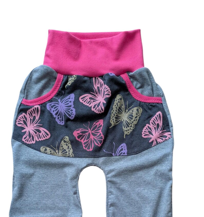 May include: Gray baby pants with a pink waistband and pocket trim. The front panel features a dark gray fabric with pink, purple, and gold butterfly designs. The pants have a high waist and a comfortable fit.