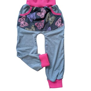 May include: Gray baby pants with a pink waistband and cuffs. The front features a dark gray panel with pink, purple, and gold butterfly designs. The pants have a high waist and a comfortable, relaxed fit.