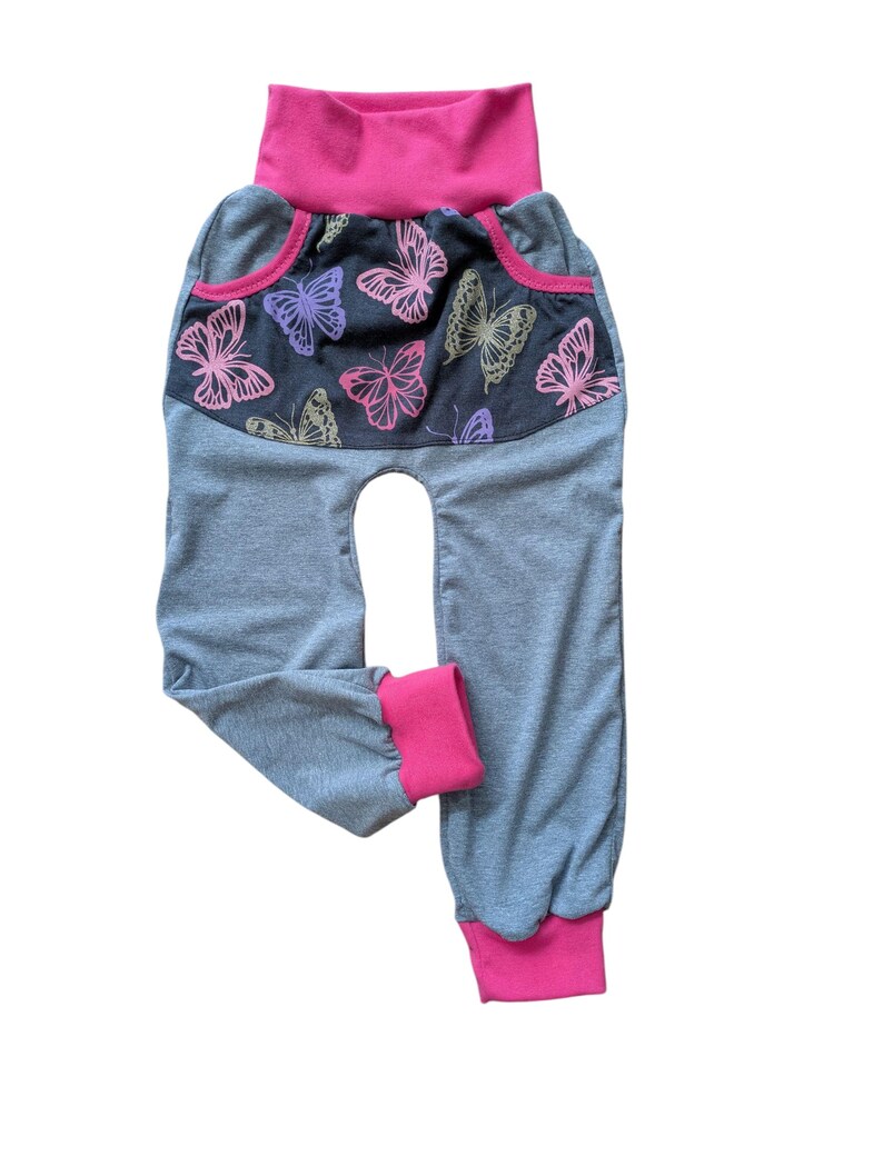 May include: Gray baby pants with a pink waistband and cuffs. The front features a dark gray panel with pink, purple, and gold butterfly designs. The pants have a high waist and a comfortable, relaxed fit.