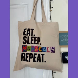 May include: A beige canvas tote bag with the words "EAT. SLEEP. MUSICALS. REPEAT." printed in black and colourful letters. The bag is hanging on a white door. The bag has two handles.