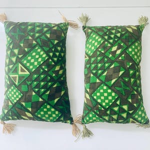 May include: Two rectangular cushions with a geometric pattern in shades of green and brown. Each cushion has a tassel at each corner. The pattern includes squares, triangles, and diamond shapes.