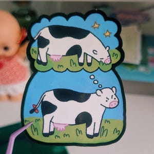 May include: A whimsical sticker featuring a cartoon cow in a blue and green scene. The cow is black and white with a pink udder, and a thought bubble shows a cow dreaming of stars. The sticker is held by purple tweezers.