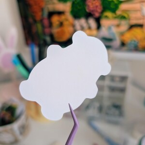 Puede incluir: Una pegatina blanca troquelada en forma de nube es sostenida por unas pinzas moradas. La pegatina es el foco principal, con un fondo borroso de objetos coloridos y una pintura. Las pinzas est&aacute;n orientadas hacia el espectador.