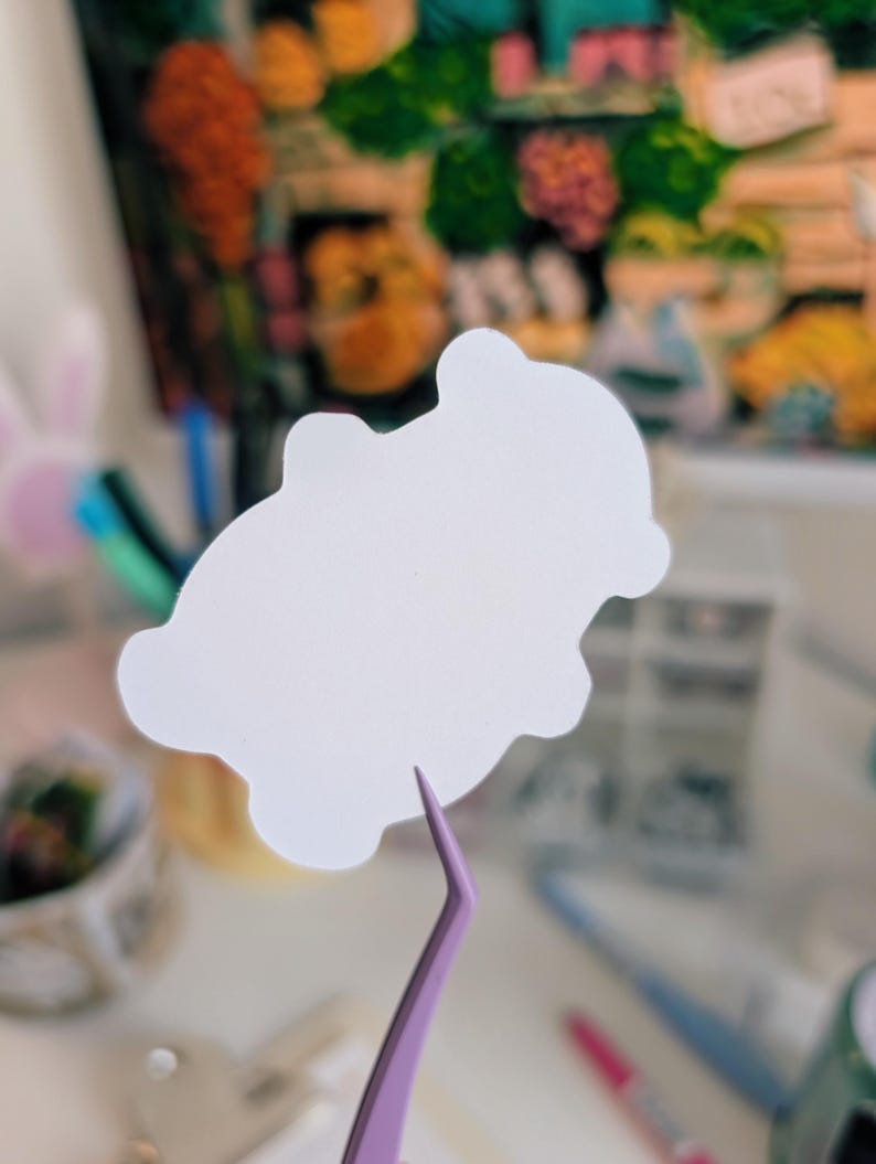 Puede incluir: Una pegatina blanca troquelada en forma de nube es sostenida por unas pinzas moradas. La pegatina es el foco principal, con un fondo borroso de objetos coloridos y una pintura. Las pinzas est&aacute;n orientadas hacia el espectador.