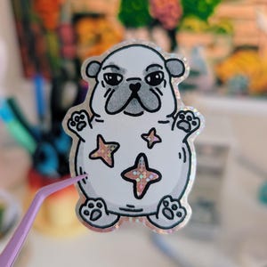 Puede incluir: Una pegatina blanca con un pug de dibujos animados con una expresi&oacute;n gru&ntilde;ona. La pegatina tiene un borde hologr&aacute;fico y detalles en forma de estrella en su cuerpo. El pug tiene contornos negros y un hocico gris. Unas pinzas moradas sostienen la pegatina.