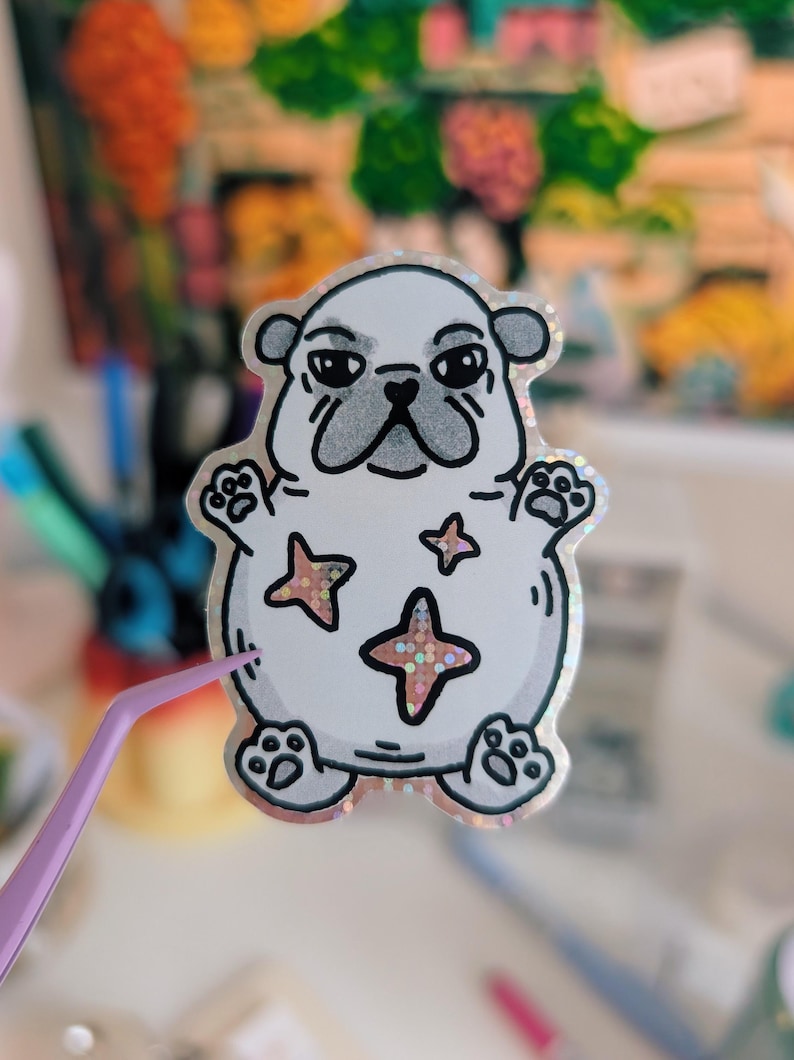 Puede incluir: Una pegatina blanca con un pug de dibujos animados con una expresi&oacute;n gru&ntilde;ona. La pegatina tiene un borde hologr&aacute;fico y detalles en forma de estrella en su cuerpo. El pug tiene contornos negros y un hocico gris. Unas pinzas moradas sostienen la pegatina.