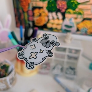 Puede incluir: Una pegatina gris claro de un perro pug de dibujos animados con estrellas. El pug est&aacute; tumbado boca arriba, sostenido por unas pinzas moradas. Una pintura colorida est&aacute; en el fondo. Otros art&iacute;culos de papeler&iacute;a tambi&eacute;n son visibles.