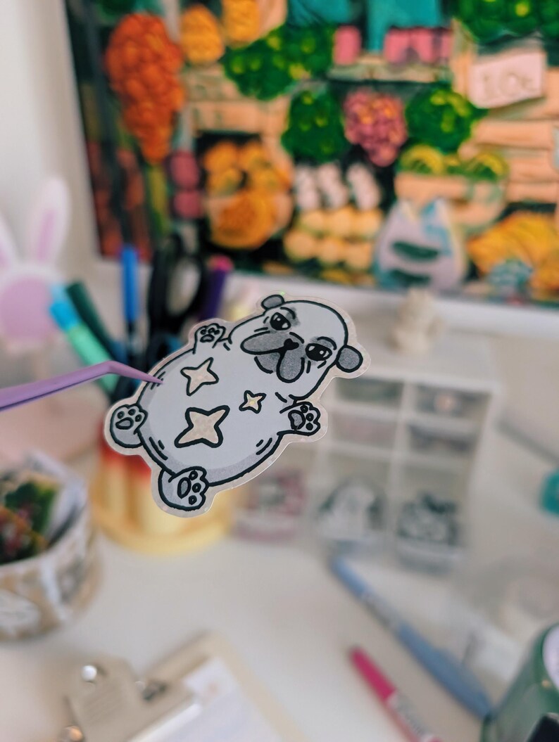 Puede incluir: Una pegatina gris claro de un perro pug de dibujos animados con estrellas. El pug est&aacute; tumbado boca arriba, sostenido por unas pinzas moradas. Una pintura colorida est&aacute; en el fondo. Otros art&iacute;culos de papeler&iacute;a tambi&eacute;n son visibles.