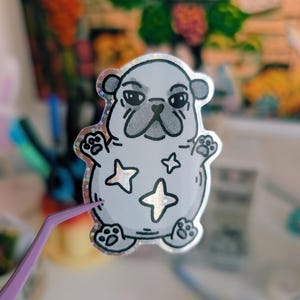 Puede incluir: Una pegatina plateada con un perro pug de dibujos animados con una expresi&oacute;n de mal humor. El pug tiene contornos negros y huellas de patas, con tres formas de estrella en su cuerpo. La pegatina tiene un borde hologr&aacute;fico y brillante.