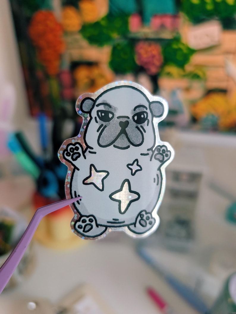 Puede incluir: Una pegatina plateada con un perro pug de dibujos animados con una expresi&oacute;n de mal humor. El pug tiene contornos negros y huellas de patas, con tres formas de estrella en su cuerpo. La pegatina tiene un borde hologr&aacute;fico y brillante.