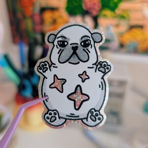 Puede incluir: Una pegatina blanca con un pug de dibujos animados con una expresi&oacute;n de mal humor. La pegatina tiene un borde hologr&aacute;fico y acentos en forma de estrella. El pug tiene contornos negros y un hocico gris. Unas pinzas moradas sujetan la pegatina.
