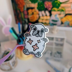 Puede incluir: Una pegatina de un pug de dibujos animados con un contorno plateado, hocico gris y tres estrellas rosas. La pegatina est&aacute; sujeta con unas pinzas moradas. El fondo incluye una pintura colorida y varios &uacute;tiles de oficina.