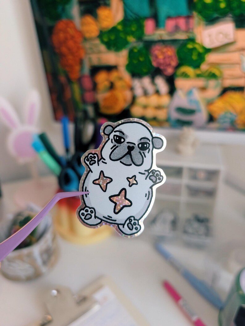 Puede incluir: Una pegatina de un pug de dibujos animados con un contorno plateado, hocico gris y tres estrellas rosas. La pegatina est&aacute; sujeta con unas pinzas moradas. El fondo incluye una pintura colorida y varios &uacute;tiles de oficina.