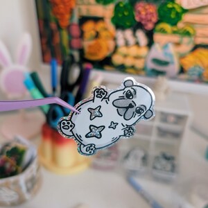 Puede incluir: Una pegatina blanca con un dise&ntilde;o de oso de dibujos animados, delineado en negro, con detalles de estrellas brillantes. La pegatina est&aacute; sujeta por unas pinzas moradas, con un fondo borroso de &uacute;tiles de arte y una pintura colorida.