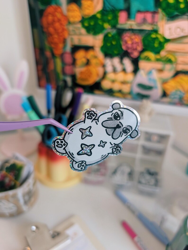 Puede incluir: Una pegatina blanca con un dise&ntilde;o de oso de dibujos animados, delineado en negro, con detalles de estrellas brillantes. La pegatina est&aacute; sujeta por unas pinzas moradas, con un fondo borroso de &uacute;tiles de arte y una pintura colorida.