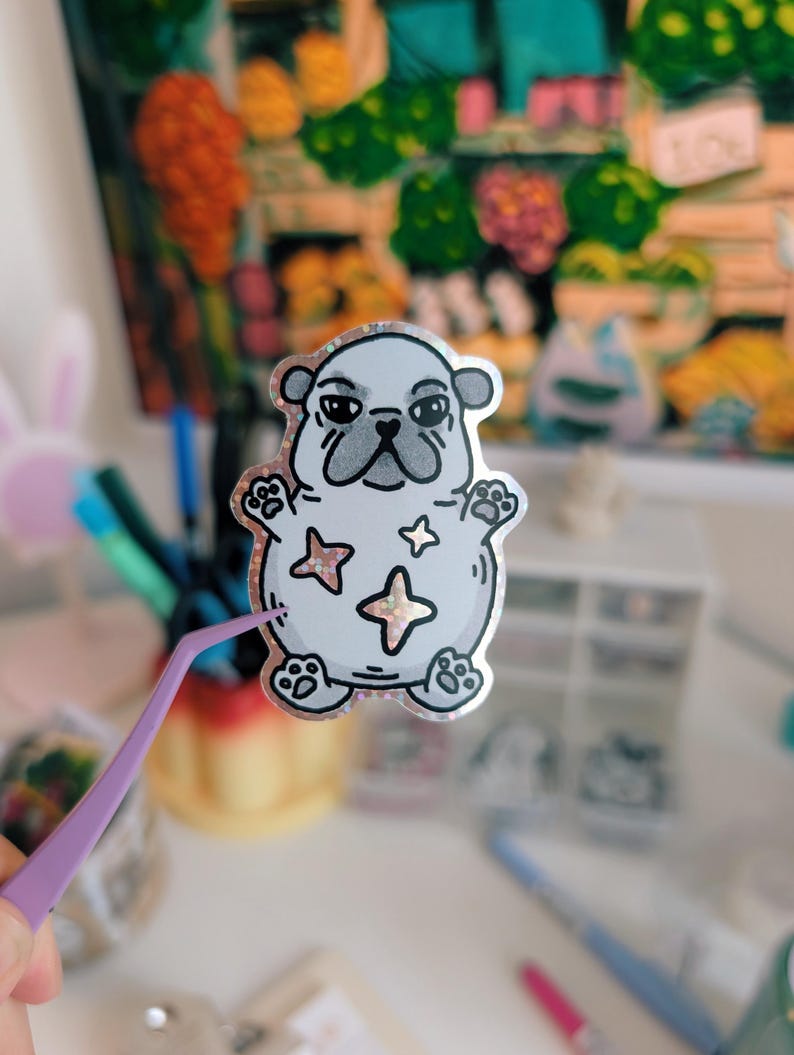 Puede incluir: Una pegatina con un dibujo de un carlino con un borde plateado brillante. El carlino es de color gris claro con rasgos faciales y patas negras. La pegatina tiene tres estrellas en rosa y dorado.