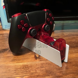 May include: A black and red PlayStation 5 controller is sitting on a white stand with the text "CRE8IVE 3D" printed on it.
