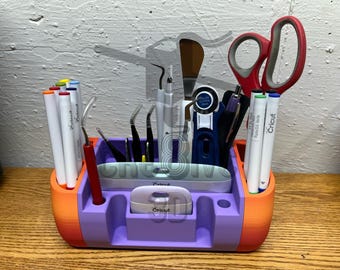 Puede incluir: Un organizador de color morado y naranja impreso en 3D con varias herramientas de manualidades, incluyendo tijeras, pinzas, bolígrafos y una herramienta Cricut. El organizador tiene una etiqueta que dice "Cricut 3D".
