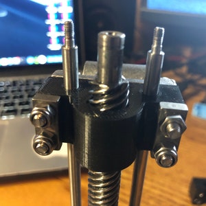 May include: A close-up of a black 3D printed part with three silver metal rods and nuts. The rods are threaded and the nuts are used to secure the rods to the part.