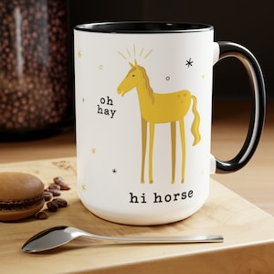 May include: White ceramic mug with a black rim and handle. The mug features a yellow cartoon horse with the text "oh hay" and "hi horse" on the front.