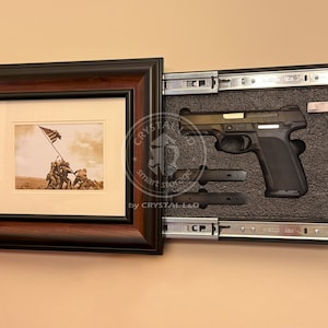 Op de afbeelding: Een donkerbruine fotolijst die een verborgen compartiment verbergt met een zwart handvuurwapen en twee magazijnen. De foto binnenin is sepia en toont soldaten die een vlag hijsen.