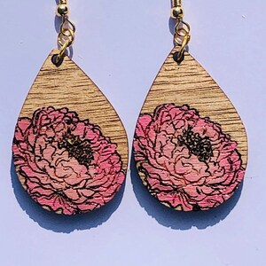 Op de afbeelding: Een paar druppelvormige houten oorbellen. Elke oorbel heeft een roze en zwart bloemenontwerp op een lichtbruine houtnerf achtergrond. De oorbellen hebben goudkleurige oorhaken.