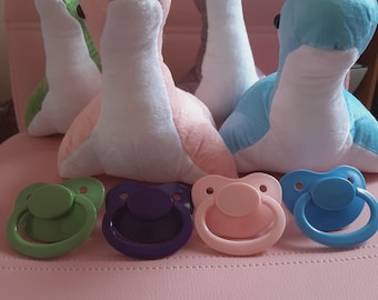 May include: A collection of plush dinosaur toys in green, pink, purple, and blue, with white bellies and necks. Below are four pacifiers in green, purple, pink, and blue. The items are arranged on a pink surface.