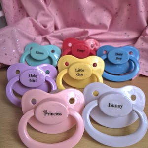 May include: A selection of colourful dummies in shades of blue, purple, pink, yellow, and red. Each dummy has a different word printed on it, including "Bunny", "Princess", and "Little One".