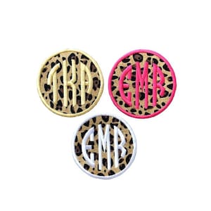 May include: Three round patches with a leopard print background. Each patch features a different monogram in a contrasting color: yellow, pink, and white. The patches are circular and have a stitched border.