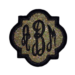 May include: A gold glitter monogram patch with black stitching in the shape of a quatrefoil. The monogram is the letters "BJM".