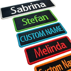 May include: Five rectangular embroidered patches with black fabric bases and stitched borders. The patches feature the names Sabrina in white, Stefan in lime green, CUSTOM NAME in cyan, Melinda in red, and a partial orange patch with orange text.