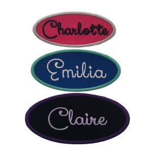 May include: Three oval-shaped embroidered patches with different names. The top patch is pink with a white outline and the name "Charlotte" in black. The middle patch is blue with a green outline and the name "Emilia" in white. The bottom patch is black with a purple outline and the name "Claire" in white.