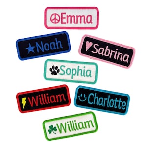 May include: Seven colorful iron-on name patches with various designs. The patches include a star, a paw print, a heart, a peace sign, a lightning bolt, a smiley face, and a shamrock. The names on the patches are Emma, Noah, Sabrina, Sophia, William, Charlotte, and William.