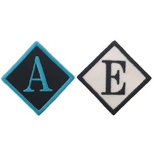 May include: Two diamond-shaped patches with embroidered letters. The first patch is black with a turquoise border and the letter "A" in turquoise. The second patch is white with a black border and the letter "E" in black.