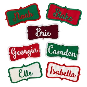 May include: Seven personalized Christmas name tags with white lettering. The tags are red, green, and white with a white border. The names on the tags are Noah, Blake, Brie, Georgia, Camden, Elle, and Isabella.