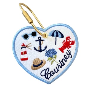 May include: Heart-shaped embroidered keychain with a light blue border and a gold-colored clasp. The white center features beach-themed designs, including a lighthouse, sunglasses, anchor, umbrella, lobster, hat, and blue flowers. The name "Courtney" is embroidered in cursive.