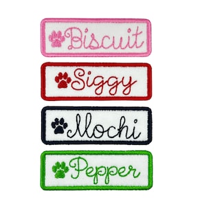 May include: Four white rectangular dog name tags with paw prints and names embroidered in different colors. The names are "Biscuit" in pink, "Siggy" in red, "Mochi" in black, and "Pepper" in green.