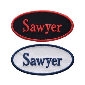 May include: Two oval-shaped embroidered patches with the word "Sawyer" in red and blue thread. The top patch is black with red lettering, and the bottom patch is white with blue lettering.