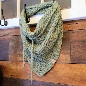 May include: A green crocheted scarf with a tassel tie hanging on a hook. The scarf is made with a simple stitch pattern and has a small brown leather tag with the word "WW" on it.