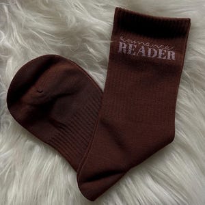 May include: A pair of brown socks with the word "READER" printed in white on the cuff. The socks are made of a soft material and are displayed on a fluffy white surface.