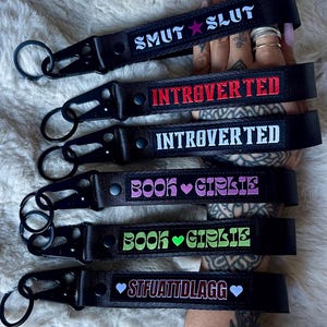 Op de afbeelding: Zwarte sleutelhangers met geborduurde tekst. De sleutelhangers tonen zinnen als "SMUT SLUT", "INTROVERTED" en "BOOK GIRLIE" in verschillende kleuren. Elke sleutelhanger heeft een metalen sluiting.