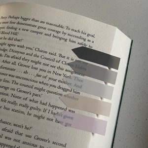 May include: A book with several arrow-shaped sticky notes in shades of gray and pink marking passages. The book is open, with text visible on the pages. The sticky notes are arranged in a vertical line.