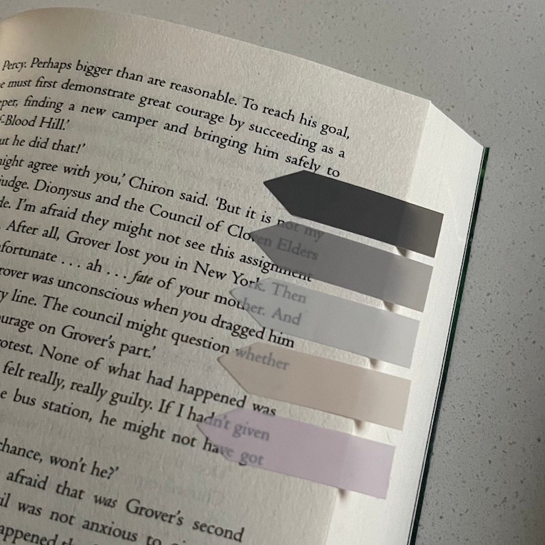 May include: A book with several arrow-shaped sticky notes in shades of gray and pink marking passages. The book is open, with text visible on the pages. The sticky notes are arranged in a vertical line.