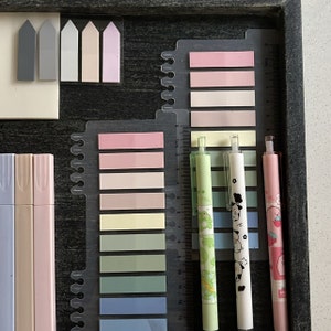 May include: A collection of stationery items including pastel-colored highlighters, sticky notes, and pens. The pens feature decorative designs, and the sticky notes are arranged in a gradient of colors. The items are neatly organized in a dark wooden tray.