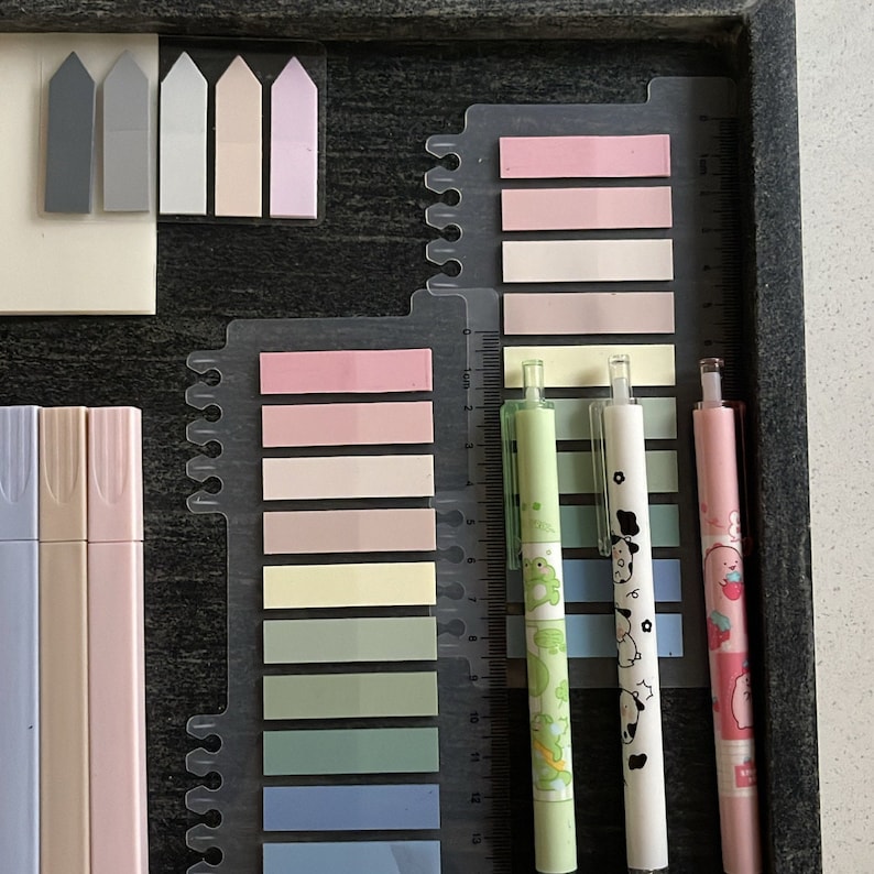 May include: A collection of stationery items including pastel-colored highlighters, sticky notes, and pens. The pens feature decorative designs, and the sticky notes are arranged in a gradient of colors. The items are neatly organized in a dark wooden tray.