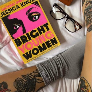 May include: A yellow book with the title "Bright Young Women" by Jessica Knoll. The book cover features a woman's face with bright pink makeup and large eyes. A pair of brown tortoiseshell glasses and a brown leather case are also in the image.