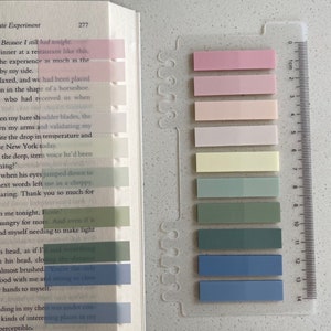 May include: A book with colorful sticky notes marking pages, next to a clear plastic ruler. The sticky notes are in shades of pink, yellow, green, and blue. The book is open to a page with text.