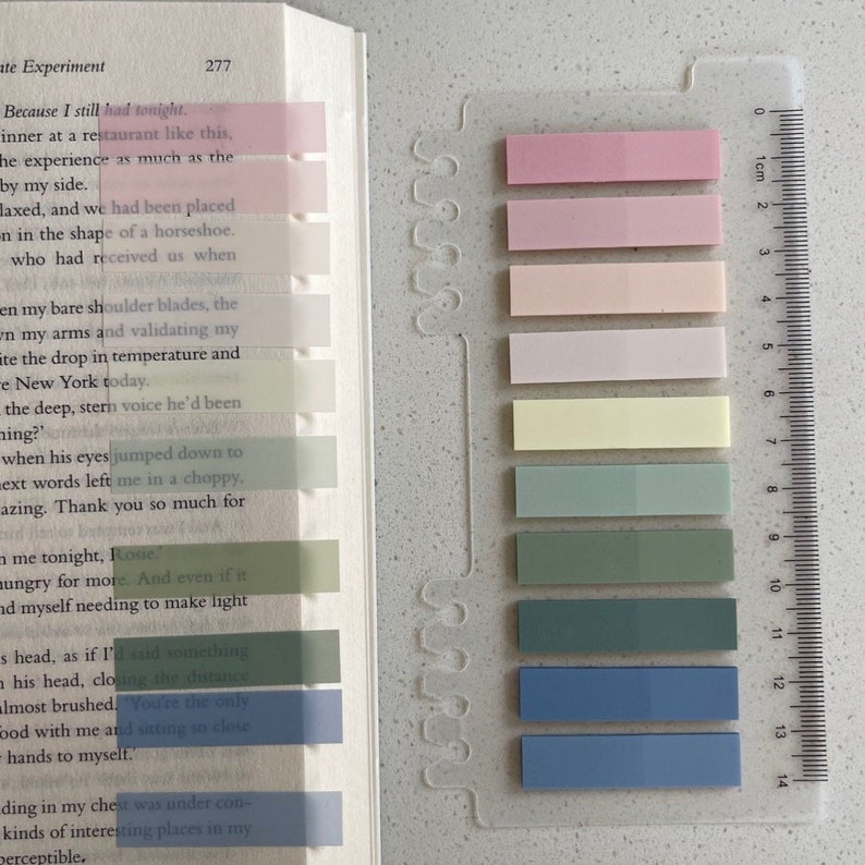May include: A book with colorful sticky notes marking pages, next to a clear plastic ruler. The sticky notes are in shades of pink, yellow, green, and blue. The book is open to a page with text.